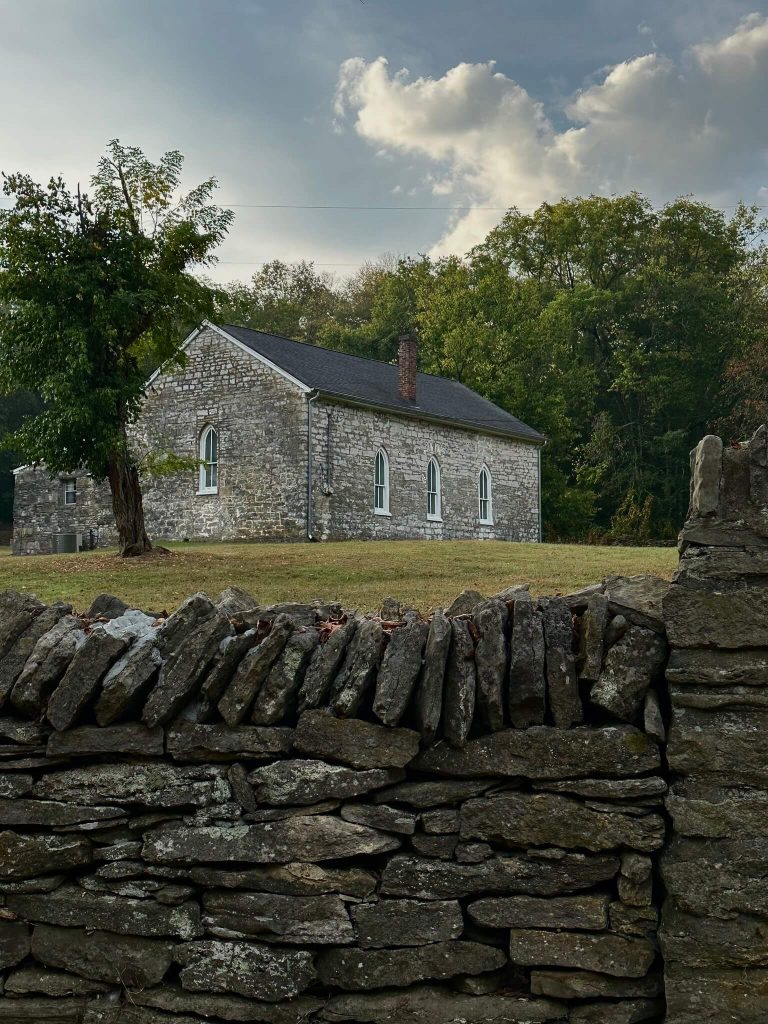 Historic limestone church with arched windows behind a dry-stacked stone fence, set against leafy trees and a cloudy evening sky.