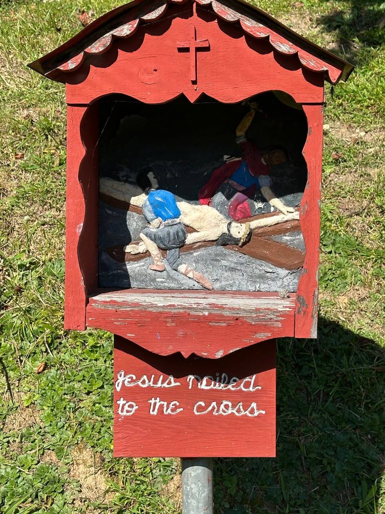 Red wooden roadside shrine showing a carved scene of Jesus being nailed to the cross, with the words “Jesus nailed to the cross.”
