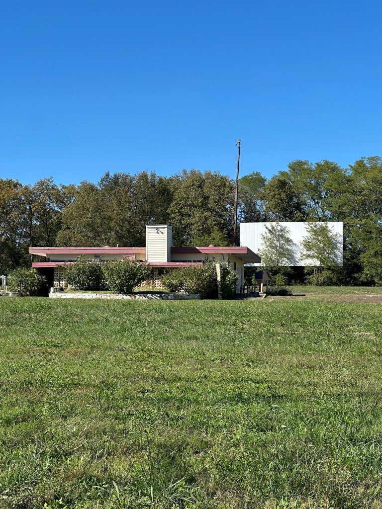 Abandoned Sky Vue Twin Drive-In concession building with overgrown shrubs and a faded screen behind, in a grassy field.