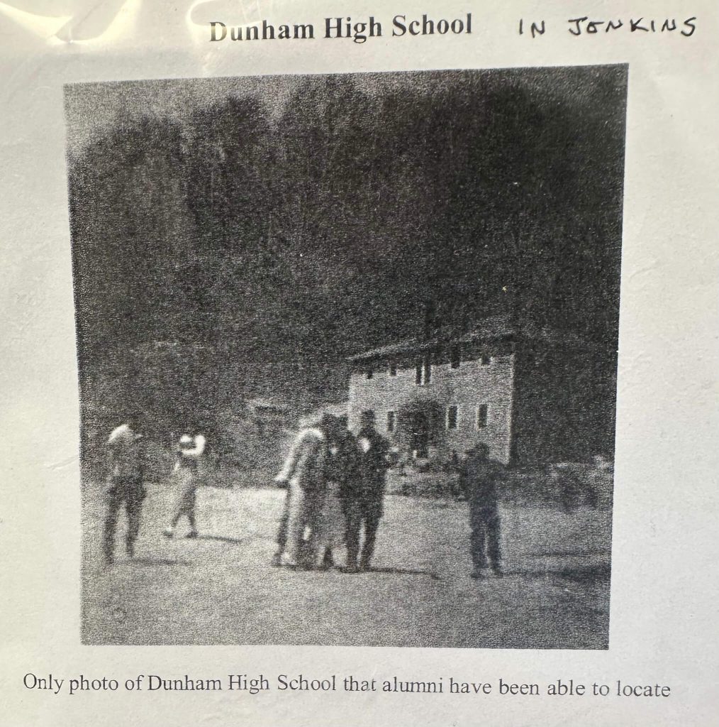 Grainy black-and-white photo labeled “Dunham High School,” showing a group of people in a field with the school building in the background against a wooded hillside.