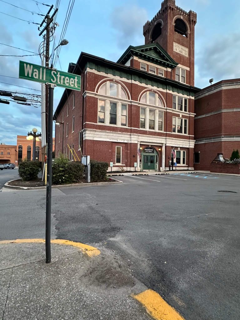 Winchester City Hall on Wall Street at dusk, showing the tall brick clock tower and arched windows beside an empty parking lot.