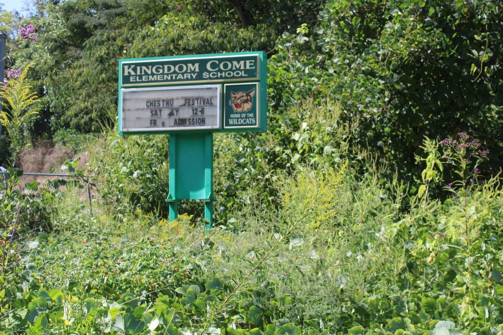 Overgrown marquee reading “Kingdom Come Elementary School,” with a wildcat logo and a faded community notice board surrounded by tall weeds and trees.