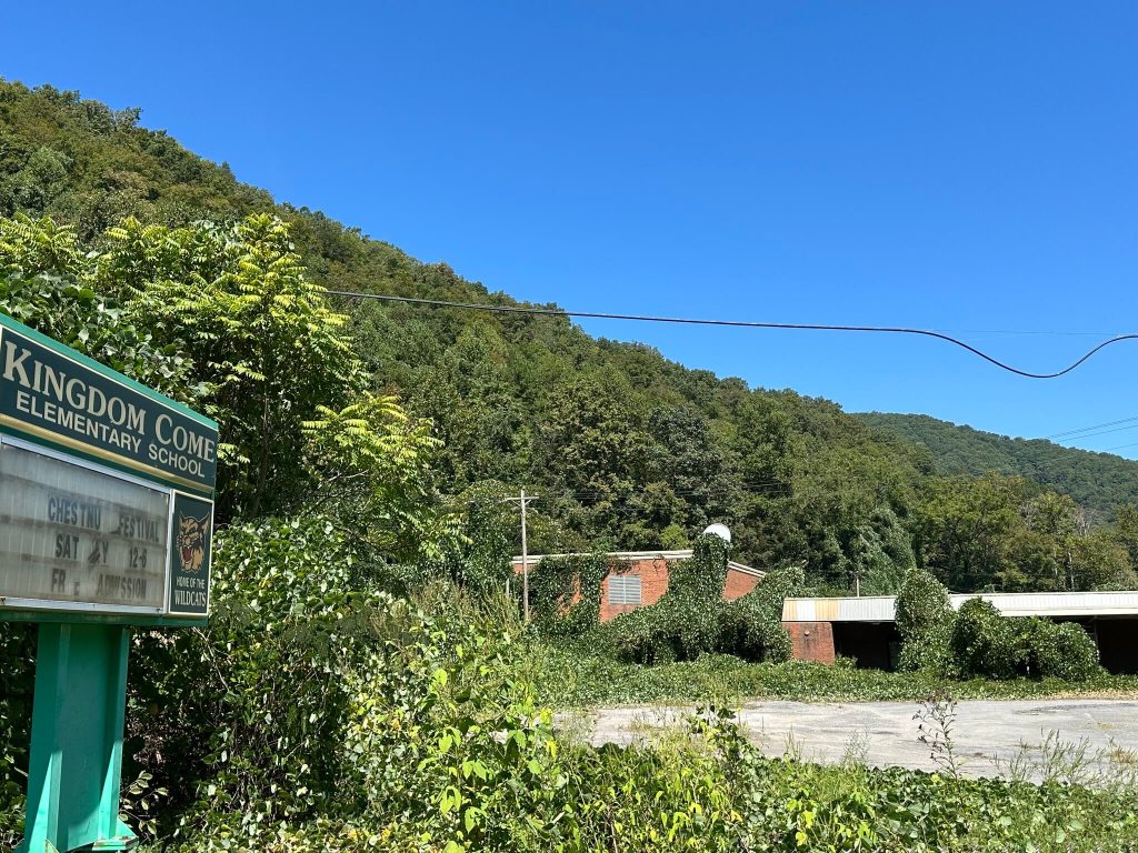 Kingdom Come Elementary School sign at left, with the abandoned, vine covered brick school buildings and empty lot beneath forested mountains in Linefork, Kentucky.