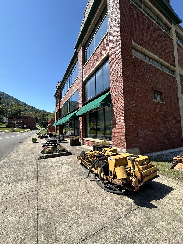 Long sidewalk view beside the Kentucky Coal Museum in Benham, Kentucky, featuring the tall red brick company store with green awnings, historic yellow mining equipment, picnic tables, flower beds, and the mountains rising in the background.