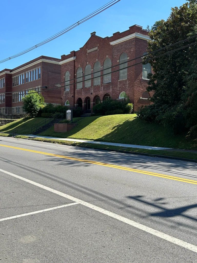 Red brick Jenkins United Methodist Church with tall arched windows beside the old Jenkins school, viewed from across Main Street with a grassy slope and steps in the foreground.