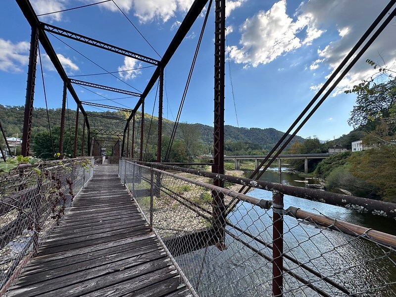 View along the old Elkhorn City walk bridge, a narrow rusted steel truss span over the Russell Fork with wooden planks and mountain hills in the background.