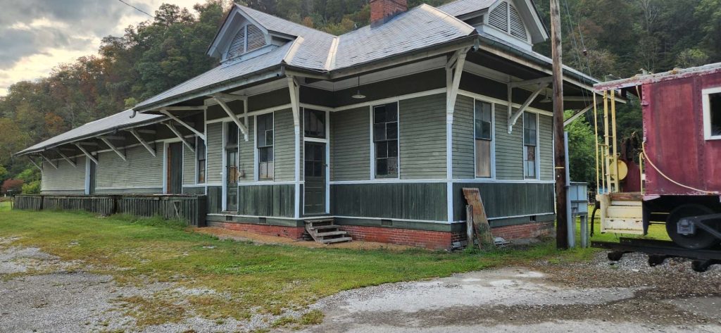 Long view of the historic green-and-gray former L&N train depot at Evarts, Kentucky, with boarded windows, brick foundation, and an overgrown railroad track stretching toward the mountains.